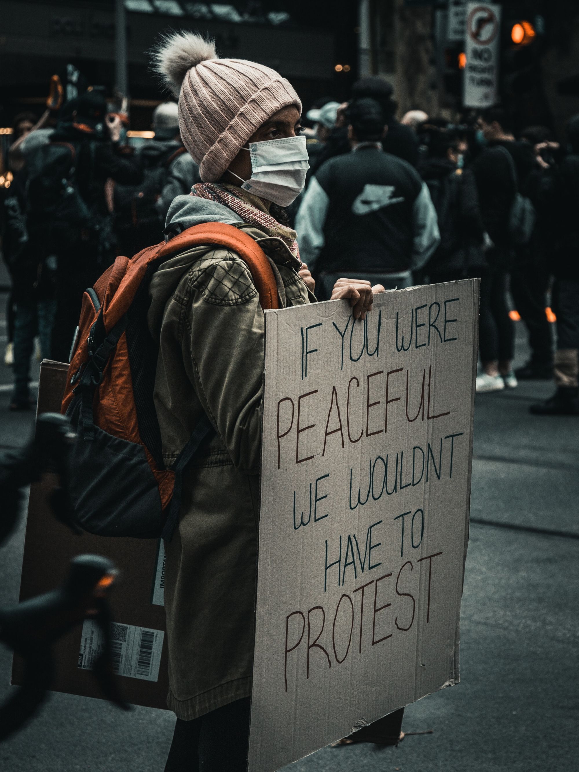 Protester in hat holding a cardboard sign "If you were peaceful, we wouldn't have to protest"